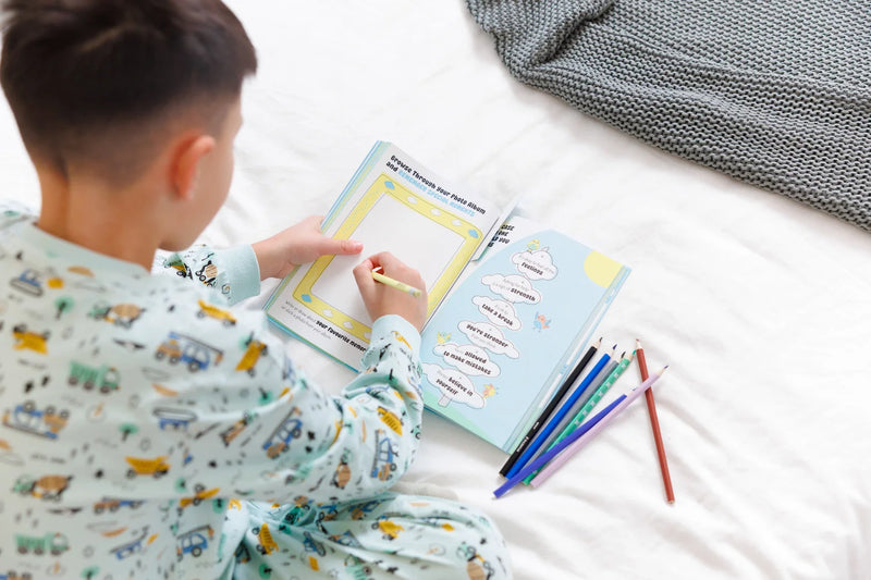 A young boy in construction-themed pajamas sitting on a bed, writing in an open InClouds. guided journal with colorful pencils nearby. The page shows positive affirmations and a space for a photo.