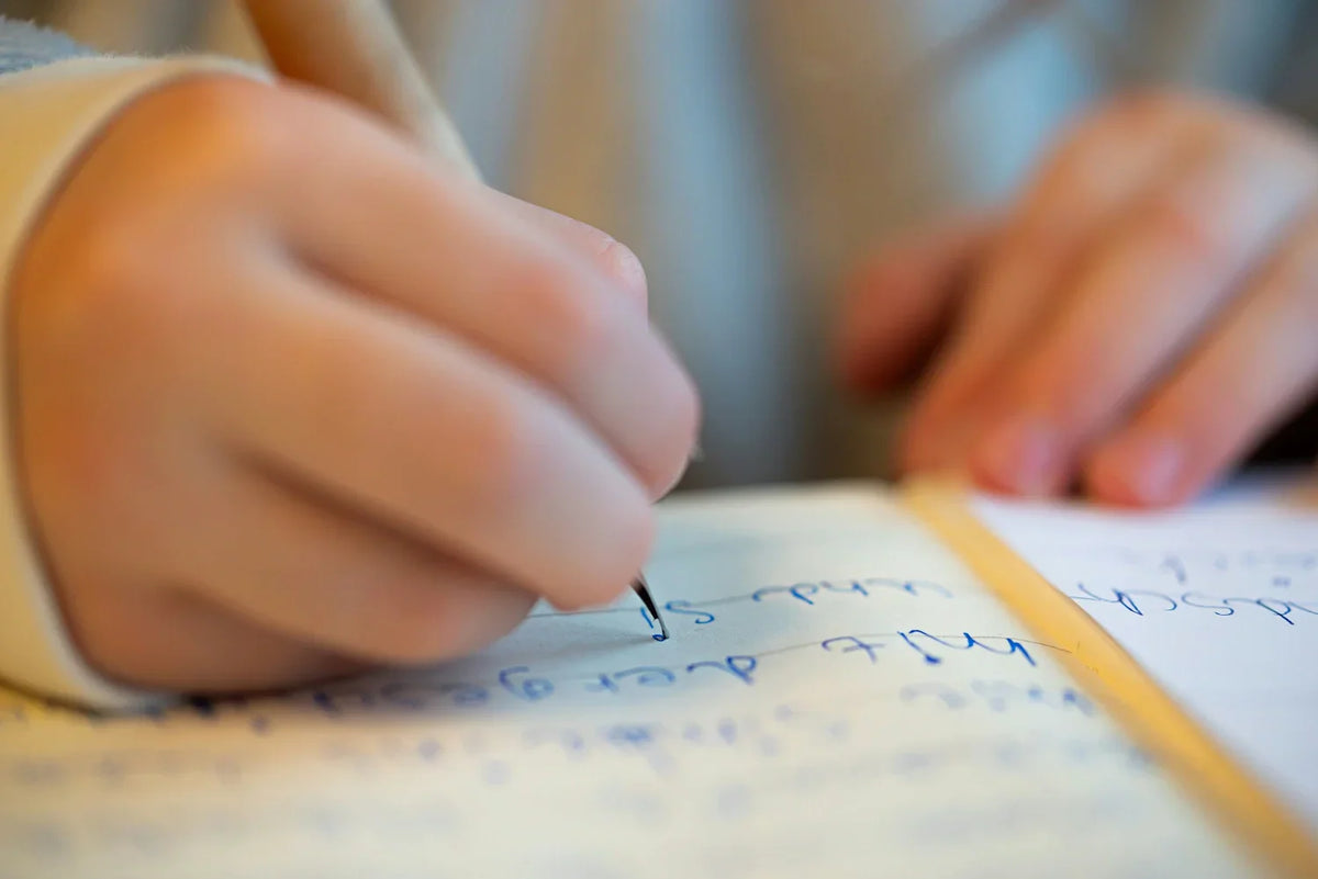 Opcija 1: Fokus na aktivnost i alat (Preporuka)  "Close-up of a child's hand writing in a journal with a pen. The image captures the focus and concentration involved in daily journaling and mindfulness practice with the InClouds Journal for kids.