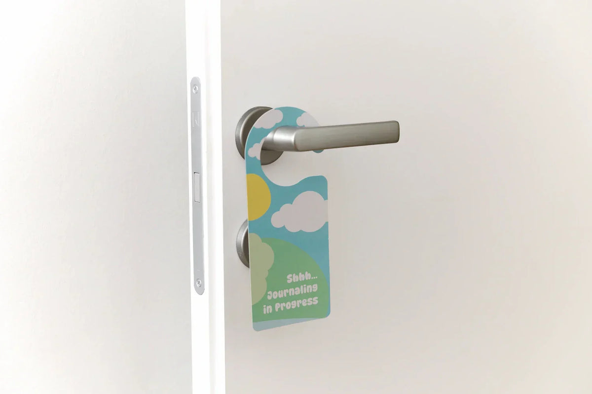 A colorful 'Shhh... Journaling in Progress' door hanger hanging on a white door handle. This InClouds Journal accessory helps children create a private space for their daily mindfulness and gratitude practice.