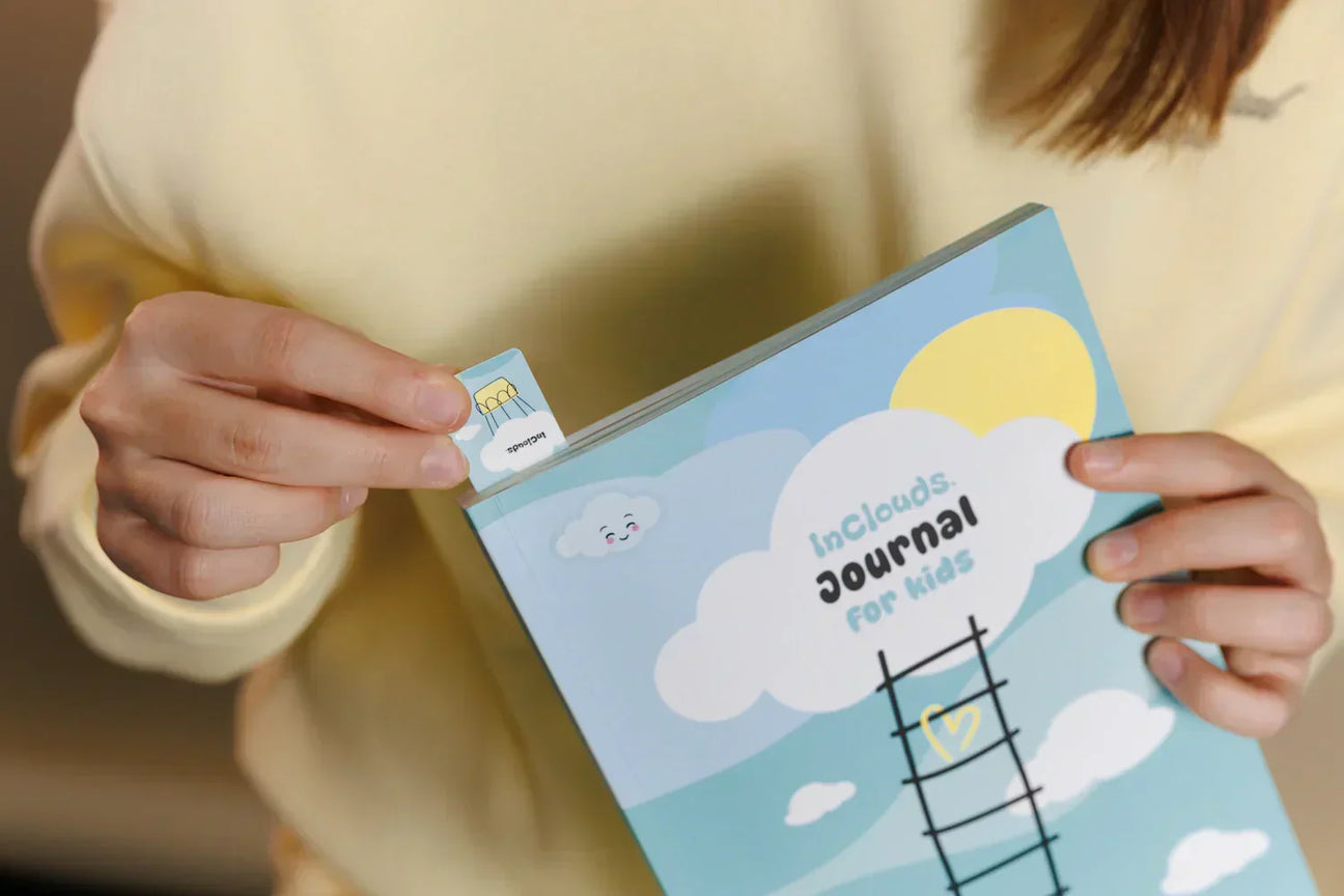 Close-up of a person holding the light blue InClouds Journal for kids and using a small illustrated bookmark. The journal cover features a ladder, clouds, and a sun, highlighting it as a high-quality mindfulness tool for children.