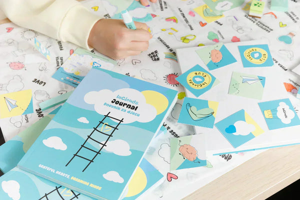 A child's hand using a marker on a creative activity mat surrounded by the InClouds Journal for kids, stickers, and cards. The turquoise gratitude journal 'Dnevnik za djecu' is the centerpiece of this mindful and educational setting.