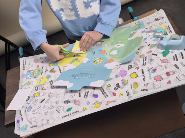 A child using green scissors to cut out colorful illustrations from a large InClouds creative activity sheet. The table is covered with stickers, tape, and branded designs, showcasing a hands-on DIY crafting experience for kids.