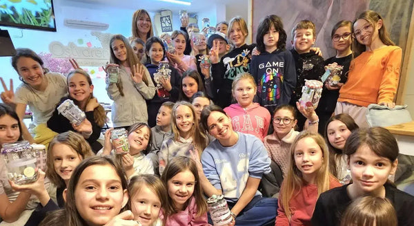 A large group of smiling children and instructors in a creative workshop holding up decorated jars.