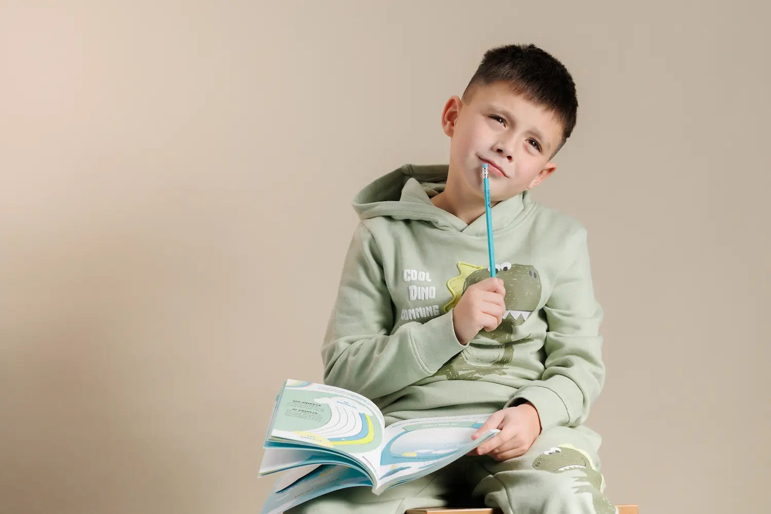 a young boy sitting against a plain beige background, holding an open workbook on his lap.
