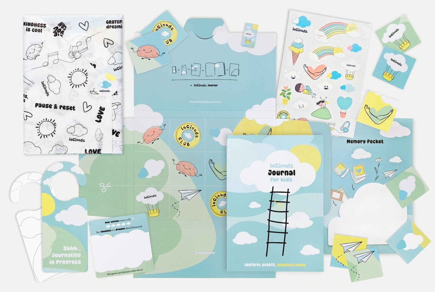 Flat lay prikaz cijelog InClouds. Journal seta dnevnika za djecu, koji uključuje vođeni dnevnik, list sa šarenim naljepnicama, mapu 'Memory Pocket' (džep za uspomene), ukrasni papir, vješalicu za vrata i male kartice s afirmacijama u nijansama plave, zelene i žute boje.