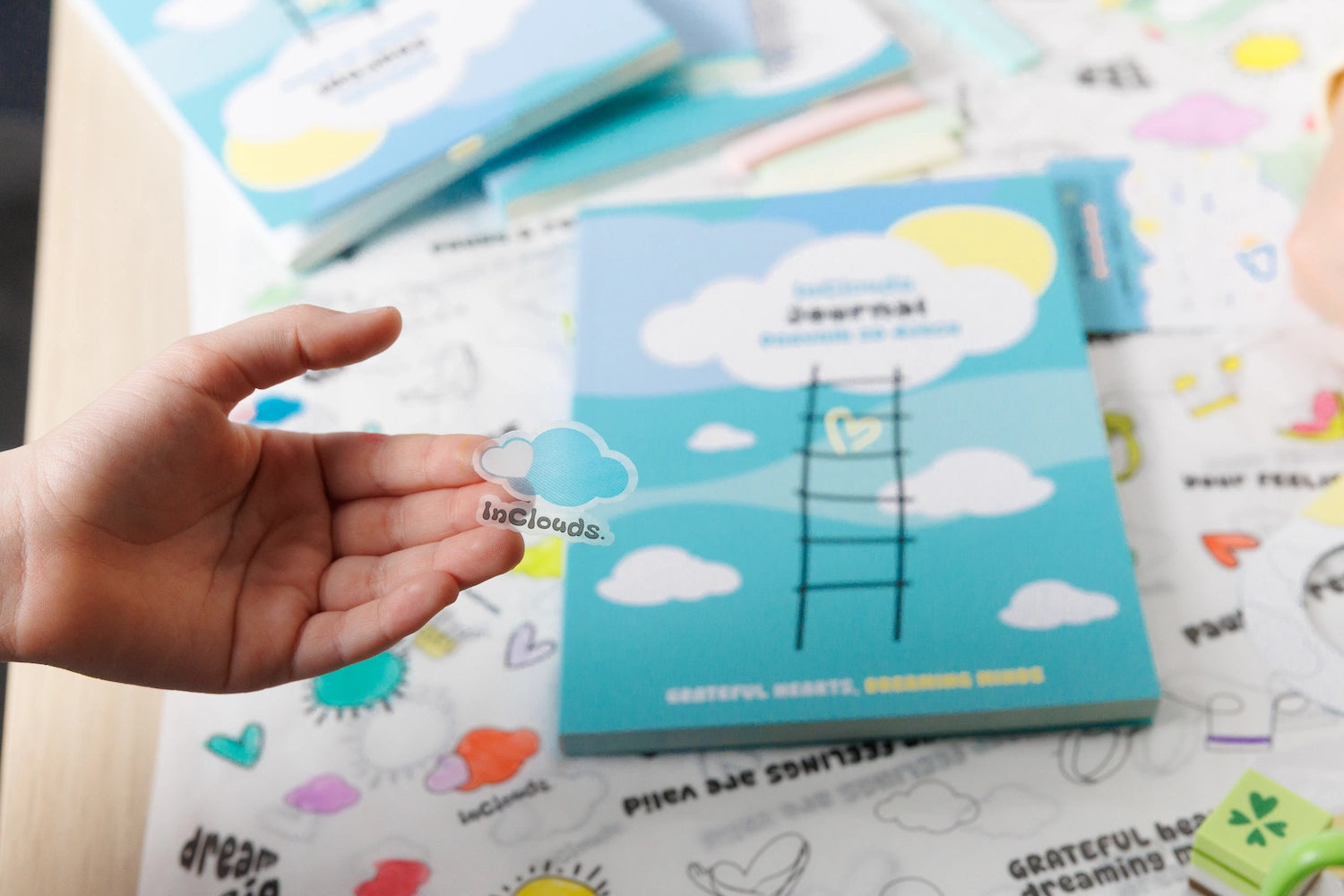 A hand holding a transparent InClouds.  cloud sticker with a blue kids journal in the background.