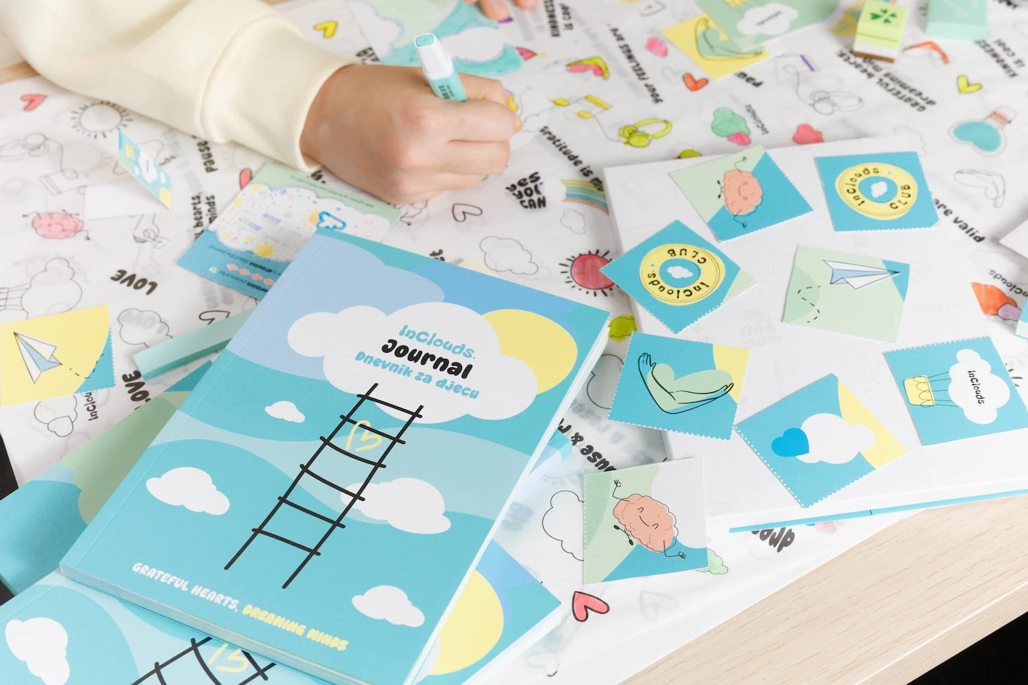 Close-up of the InClouds Journal for kids surrounded by illustrated cards and a coloring mat with a child holding a marker.